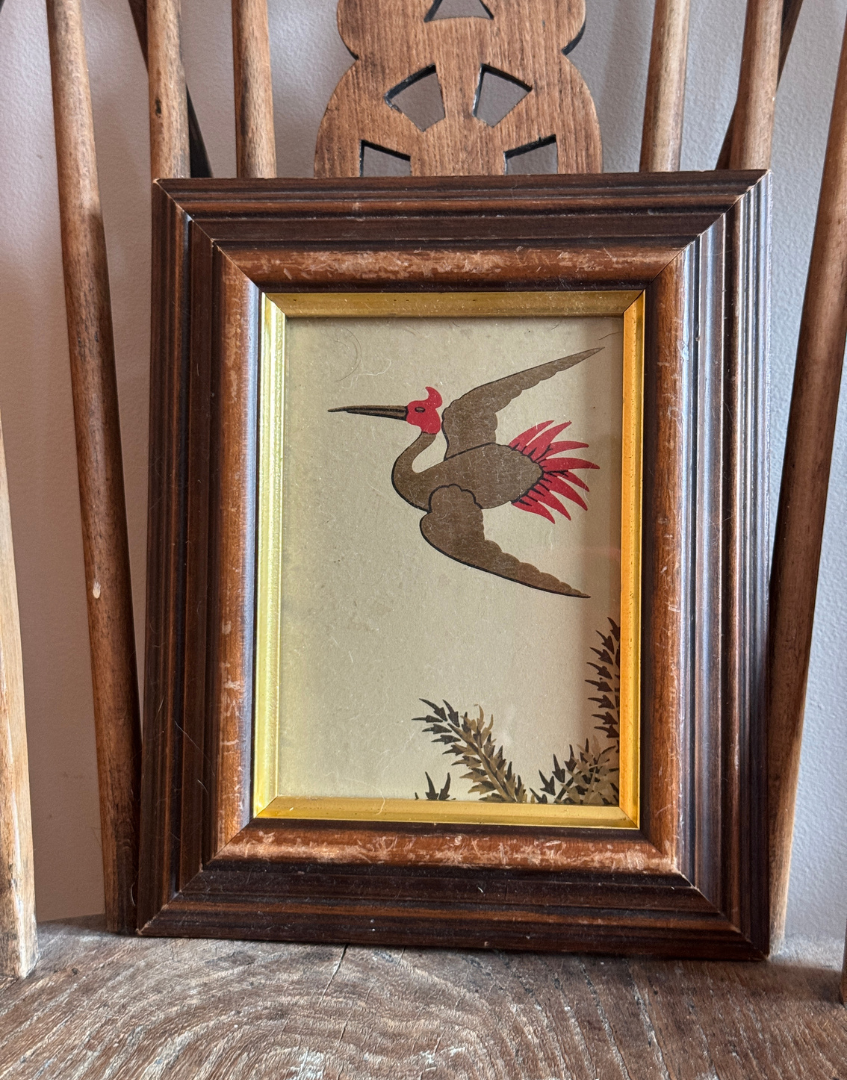 Framed artwork of a bird with red accents on a wooden chair.