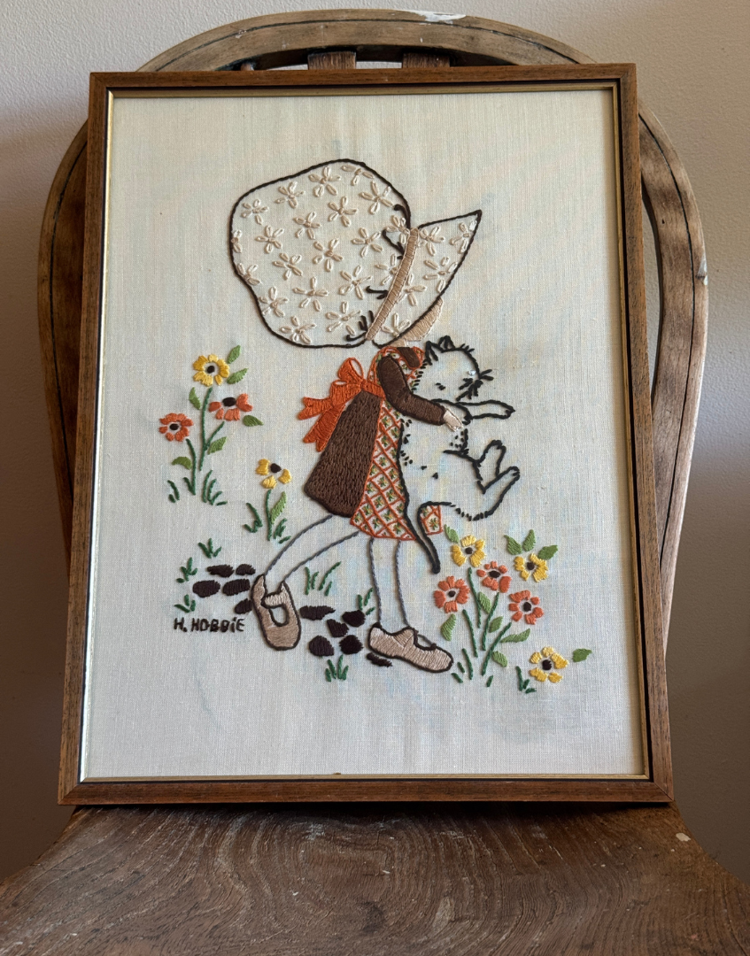 Framed Hollie Hobbie embroidery wall art in brown and gold wood frame with cream floral design.