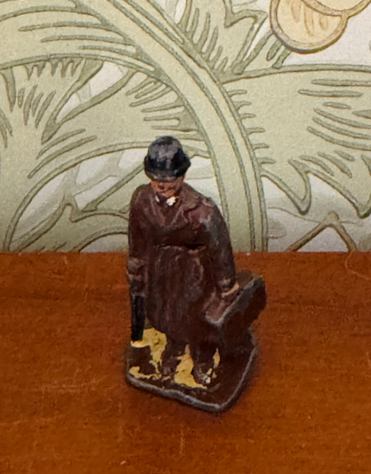 A small hand-painted miniature figurine of a man in a brown trench coat and black bowler hat, holding a suitcase, standing on a wooden surface against a patterned wallpaper background.