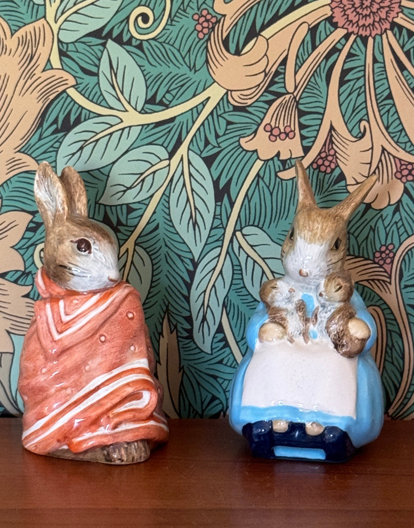 Two ceramic Peter Rabbit figurines on a wooden surface against a green and gold floral wallpaper background. The left figurine shows Peter Rabbit wrapped in an orange-red blanket, and the right figurine shows him in a blue outfit holding a white card.