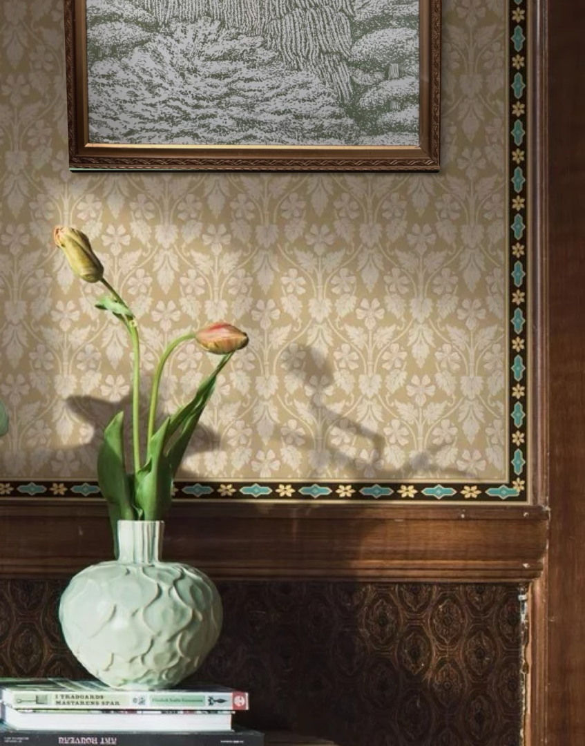 Vintage gold and wood framed classic cream and green woodland toile print in dark arts and crafts style interior with books and tulips in a white vase