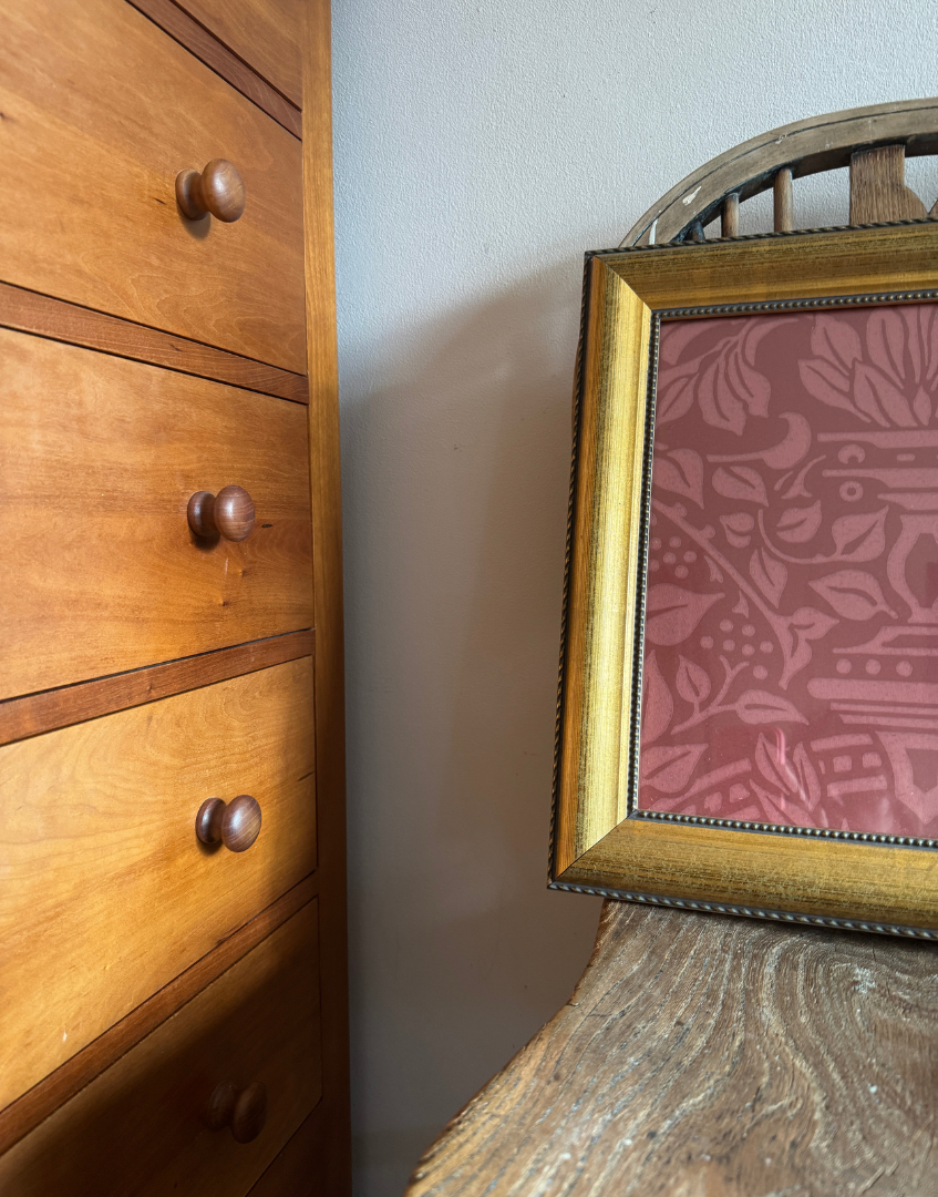 Wooden dresser with drawers next to a gold framed Gradencraft William Morris picture on an antique wooden chair seat.