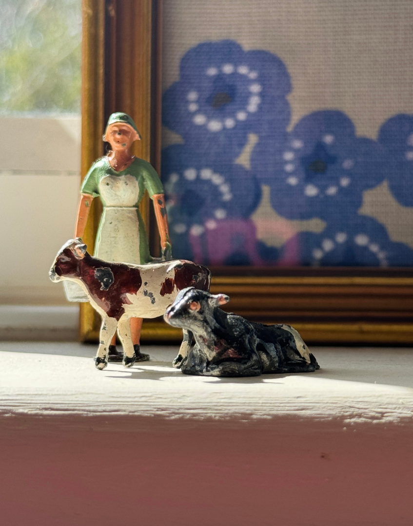 Vintage Britains lead miniature figurines featuring a milkmaid, standing calf and laying calf displayed together on a shelf.
