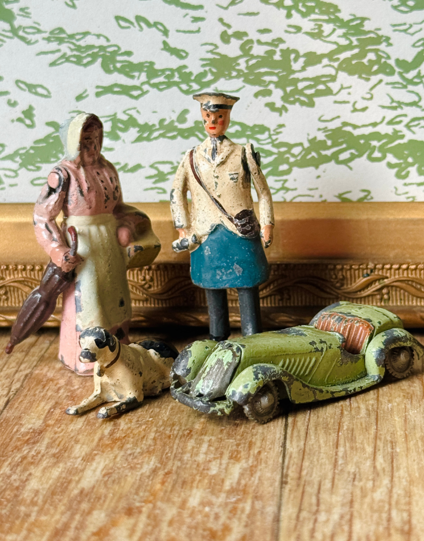 A vintage Britains toy set featuring a weathered green convertible car, a standing police officer figure, a woman holding an umbrella, and a small dog figure, arranged on a wooden surface against a patterned wallpaper background.