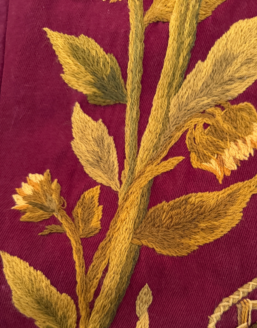 Close up of intricate crewel wool foliage embroidery with sweeping stems and leaves on the antique plum textile.