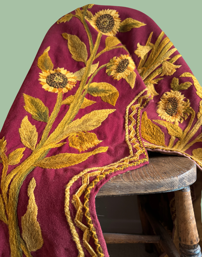 Antique Arts and Crafts crewelwork pelmet draped over an antique chair, showing its rich plum ground and large scale floral embroidery.