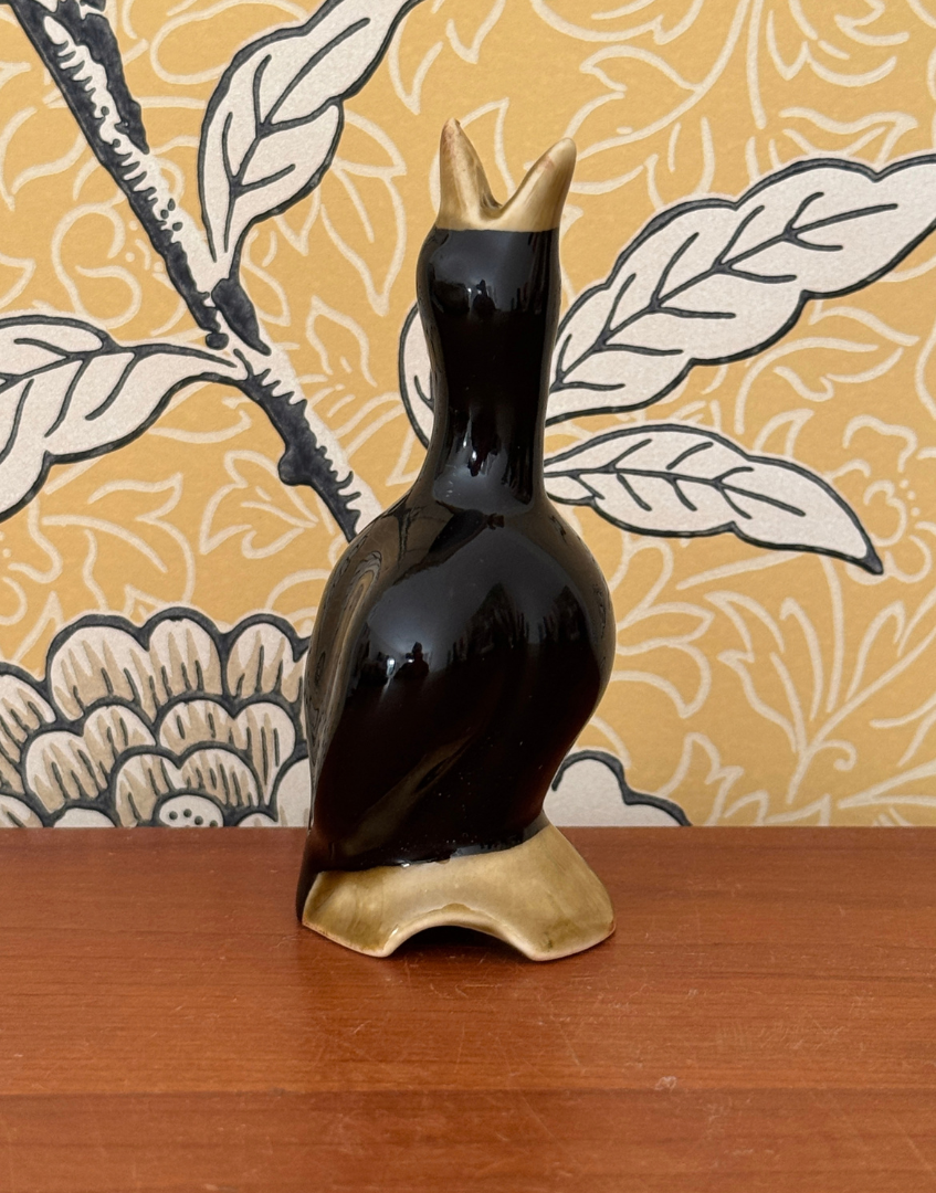 Vintage black ceramic pie bird on a wooden surface against yellow botanical wallpaper, with its open beak pointing upwards.