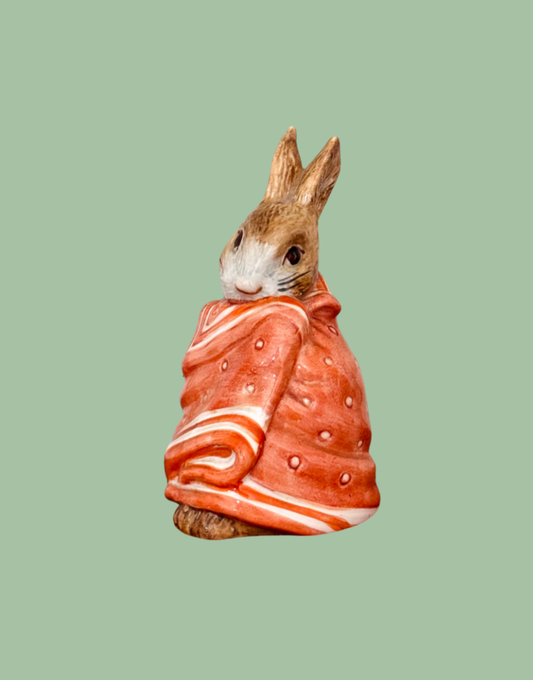 A Beswick ceramic figurine of Pooly Peter Rabbit wrapped in a red and orange patterned blanket, set against a solid sage green background.