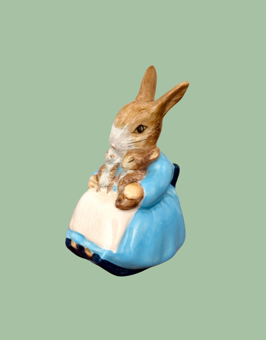 A Royol Doulton ceramic figurine of a rabbit in a blue dress holding a smaller rabbit, set against a solid sage green background.
