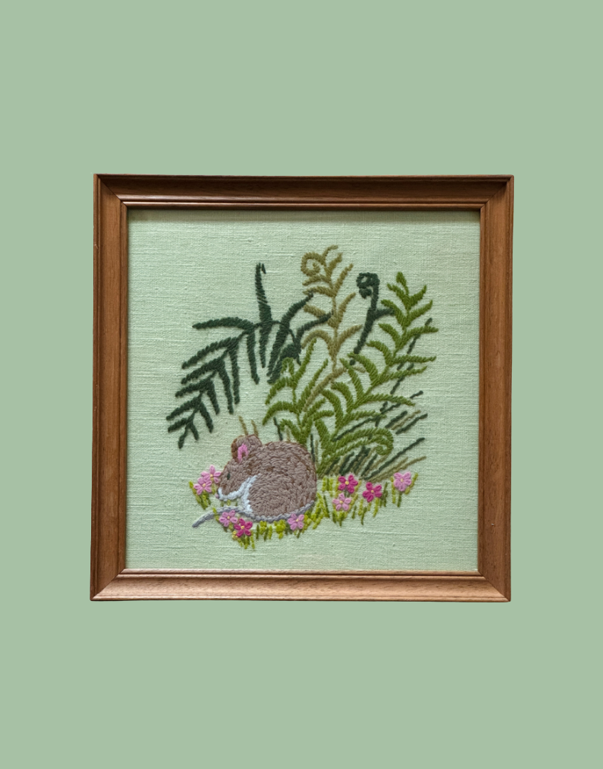A framed square needlepoint or embroidery piece featuring a brown mouse among green ferns and pink flowers on a pale green background, set in a medium brown wooden frame against a solid sage green backdrop.
