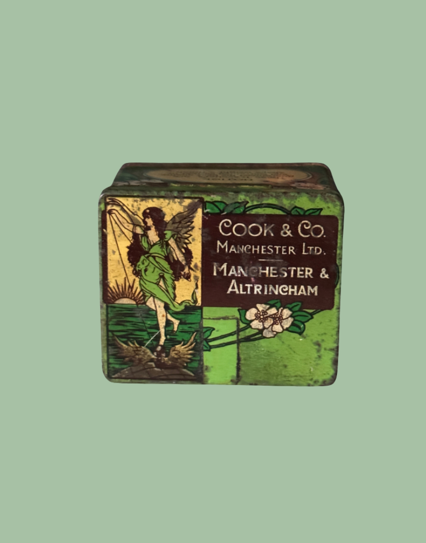 A vintage rectangular metal tin box with a painted green and brown label featuring a fairy illustration and the text 'Cook & Co Manchester Ltd. Manchester & Altringham', shown on a solid sage green background.