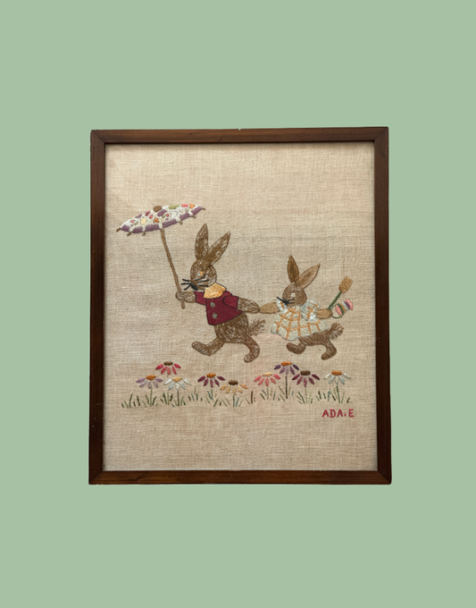 A framed vintage-style embroidered artwork showing two bunnies walking among small flowers on a beige linen background, enclosed in a dark wood frame against a pale green backdrop.
