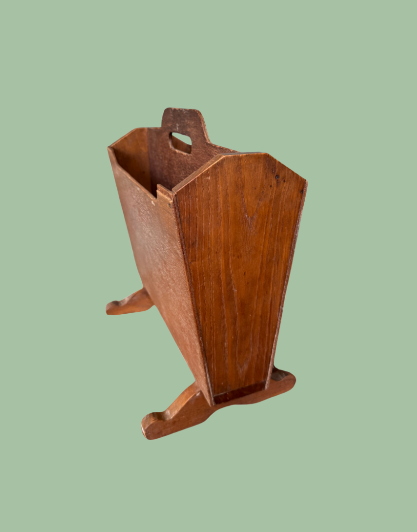 A vintage wooden magazine rack with a warm medium-brown finish, featuring a top handle cutout and small feet at the base, set against a solid sage green background.