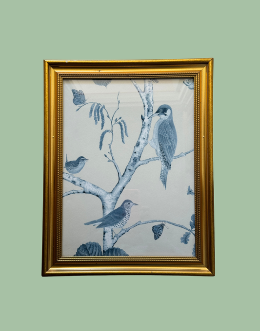 A framed art print featuring blue birds perched on birch tree branches, enclosed in a gold frame against a solid sage green background.