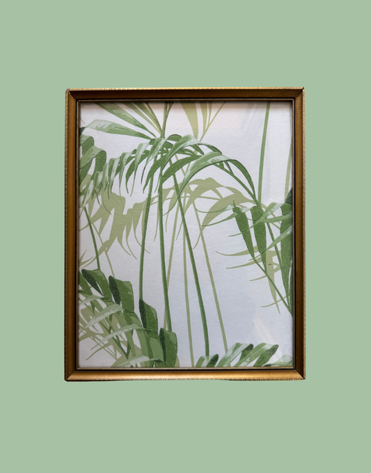 A framed botanical art print featuring green palm leaves on a white background, enclosed in a gold-toned frame against a solid sage green backdrop.