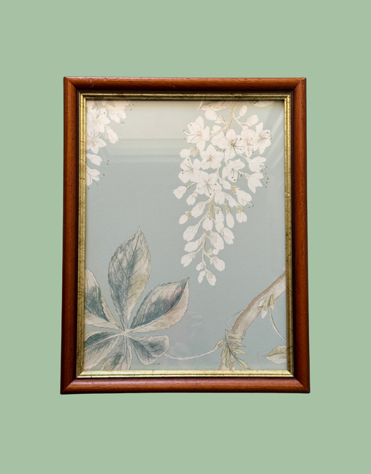 A framed botanical art print featuring white wisteria flowers and green leaves on a muted sage-gray background, enclosed in a reddish-brown wooden frame with a gold inner trim, set against a solid sage green backdrop.