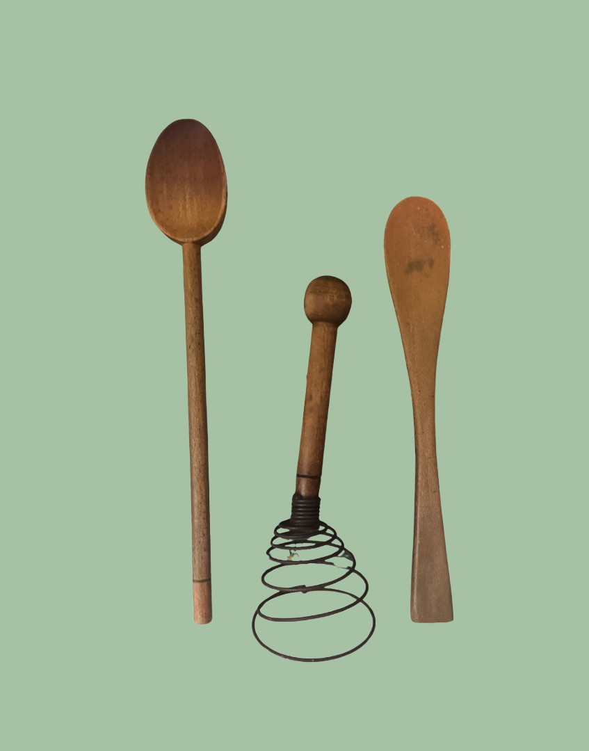 Three vintage wooden kitchen utensils, including a large spoon, a spatula, and a wooden-handled wire whisk, arranged against a solid sage green background.