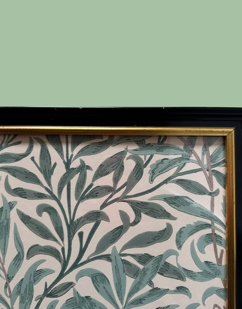 A close-up of framed botanical wallpaper featuring a repeating leaf pattern in muted teal-green on a pale blush pink background, bordered by a black frame with a gold inner trim.