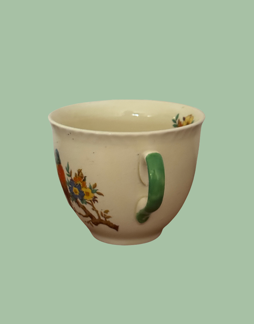 A vintage cream-colored ceramic teacup with a green handle and a painted floral design on the side, set against a solid sage green background.