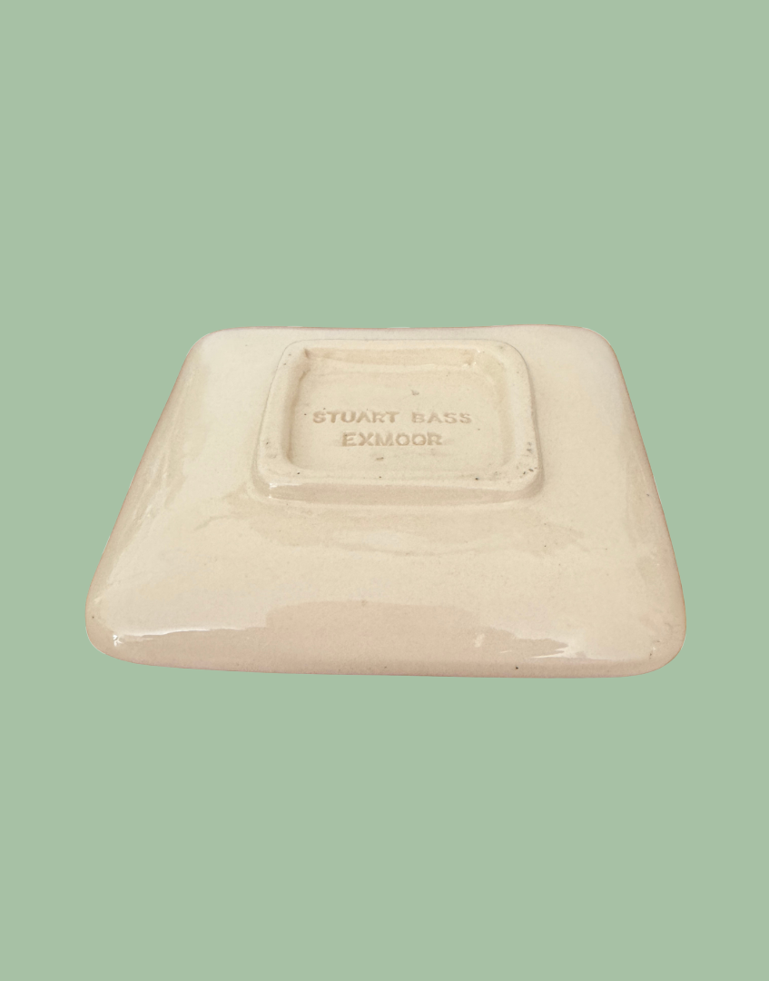 A square ceramic plate with rounded corners, shown from the back on a solid sage green background. The plate has a glossy cream-colored glaze with a stamped brand mark in the center reading 'STUART BASS EXMOOR'.
