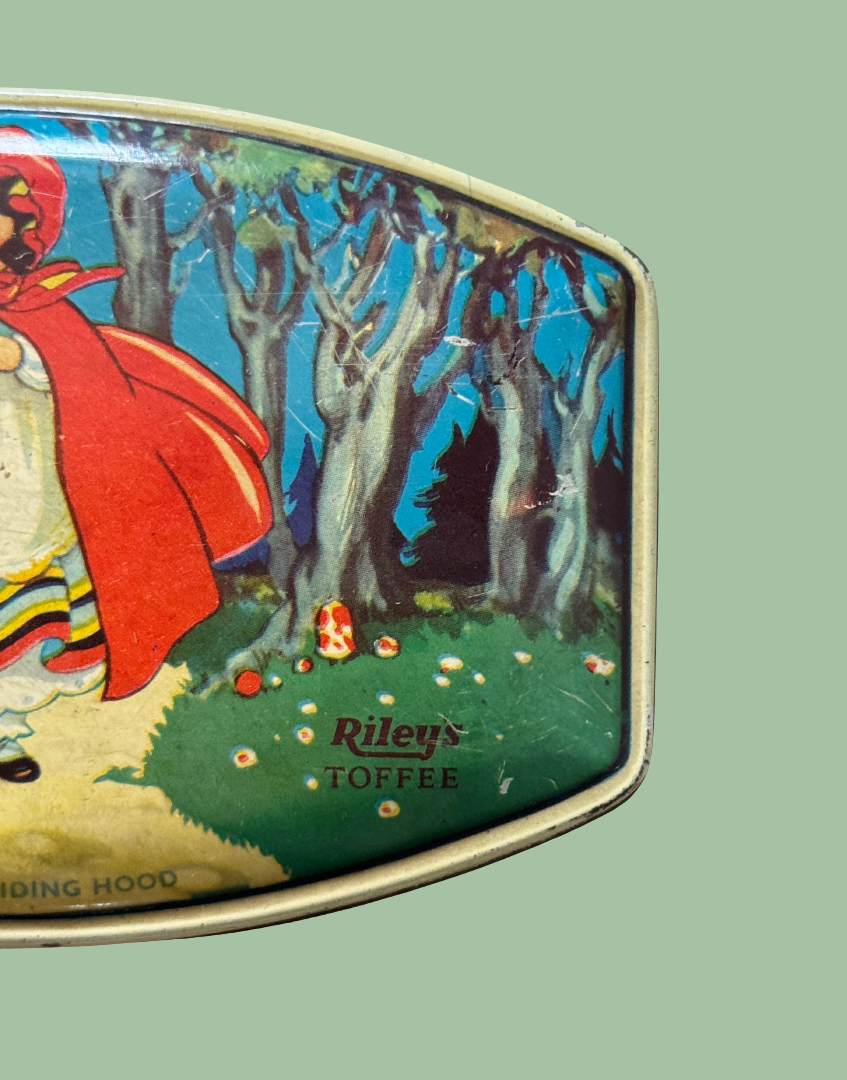 A close-up of a vintage Riley's Toffee tin with a colorful illustration of Red Riding Hood in a forest, set against a solid sage green background.