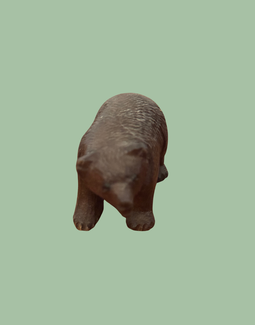 A small carved wooden bear figurine in a medium brown color, shown from a top-down perspective against a solid sage green background.