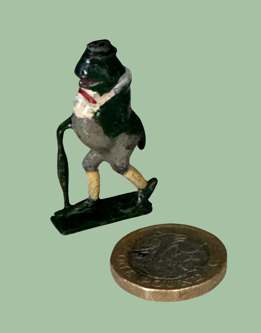 A small painted frog figurine standing on a base, shown next to a coin for scale against a plain light green background.