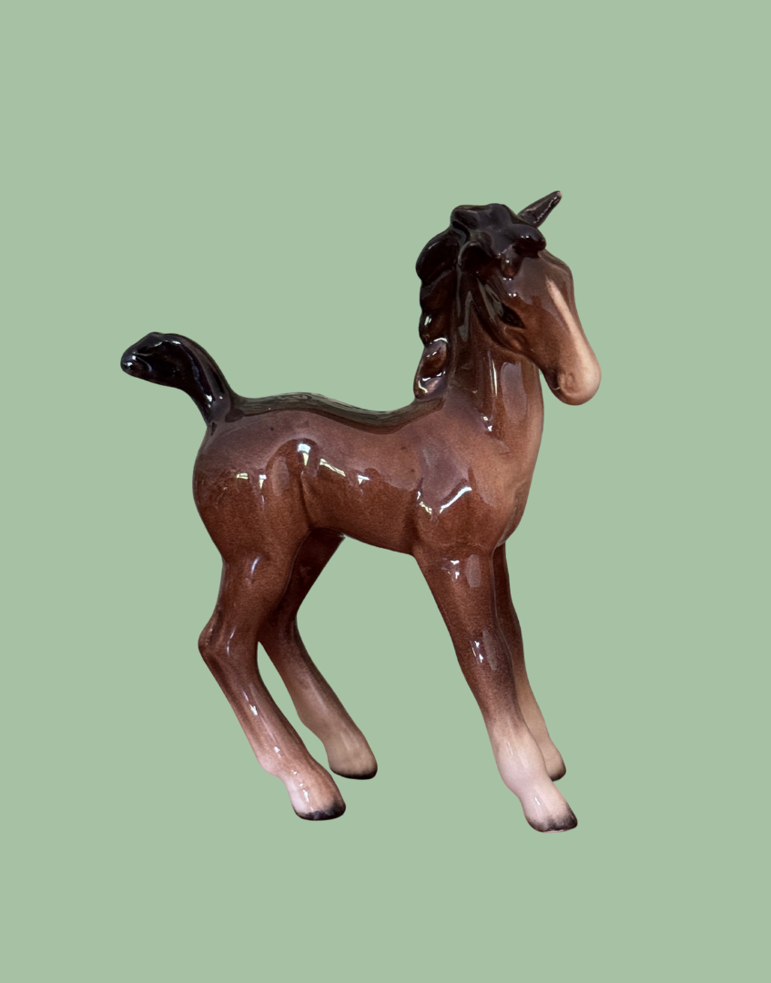 A glossy ceramic figurine of a foal horse in brown and black glaze, set against a solid sage green background.
