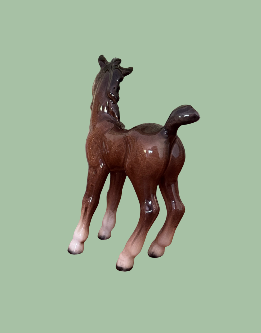 A glossy brown ceramic foal figurine with white hooves, shown from a three-quarters rear angle against a solid sage green background.