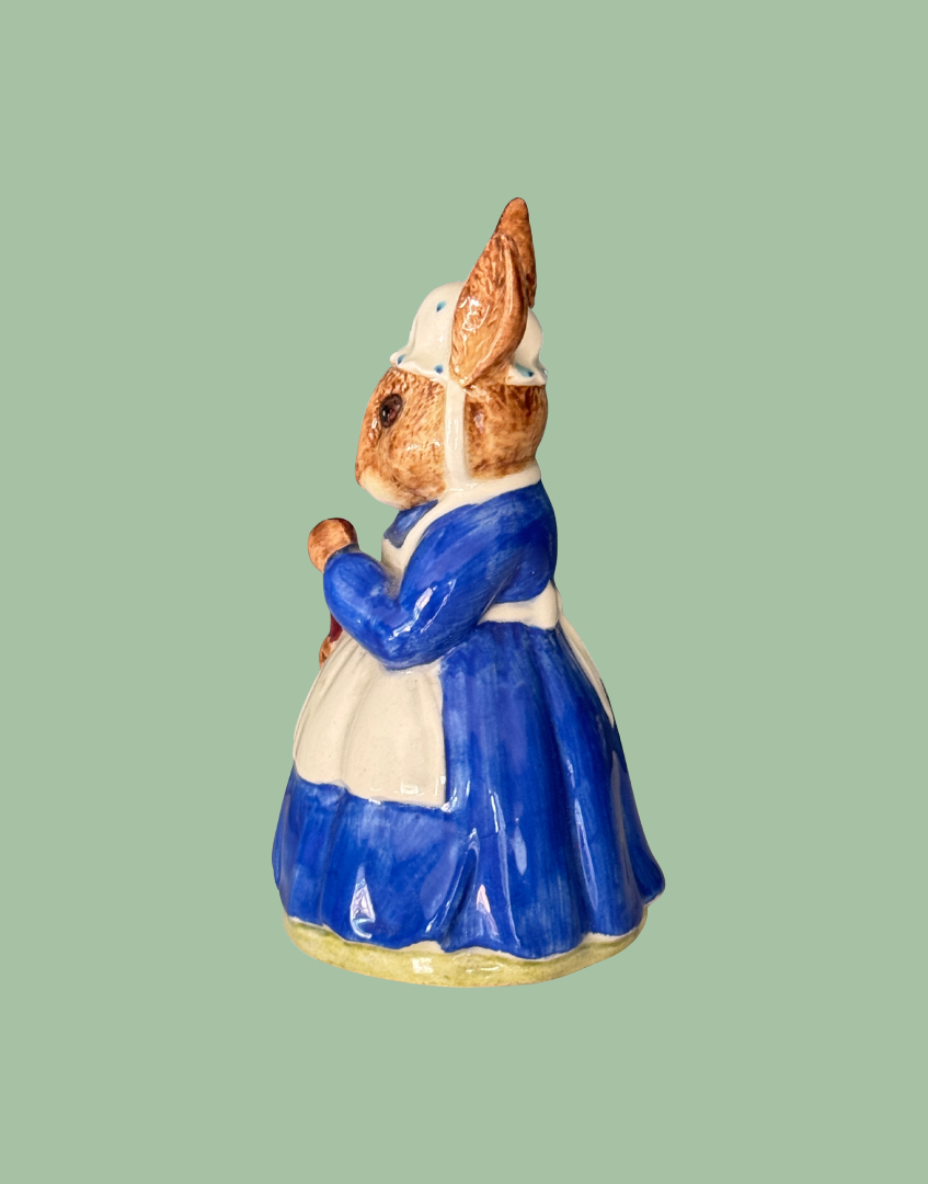 A ceramic rabbit figurine dressed in a blue dress and white apron, holding a small brown object, shown against a solid sage green background.