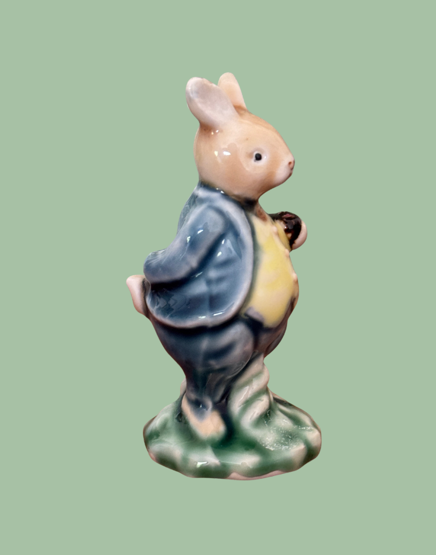A ceramic rabbit figurine wearing a blue jacket and yellow shirt, standing on a green textured base against a solid light green background.