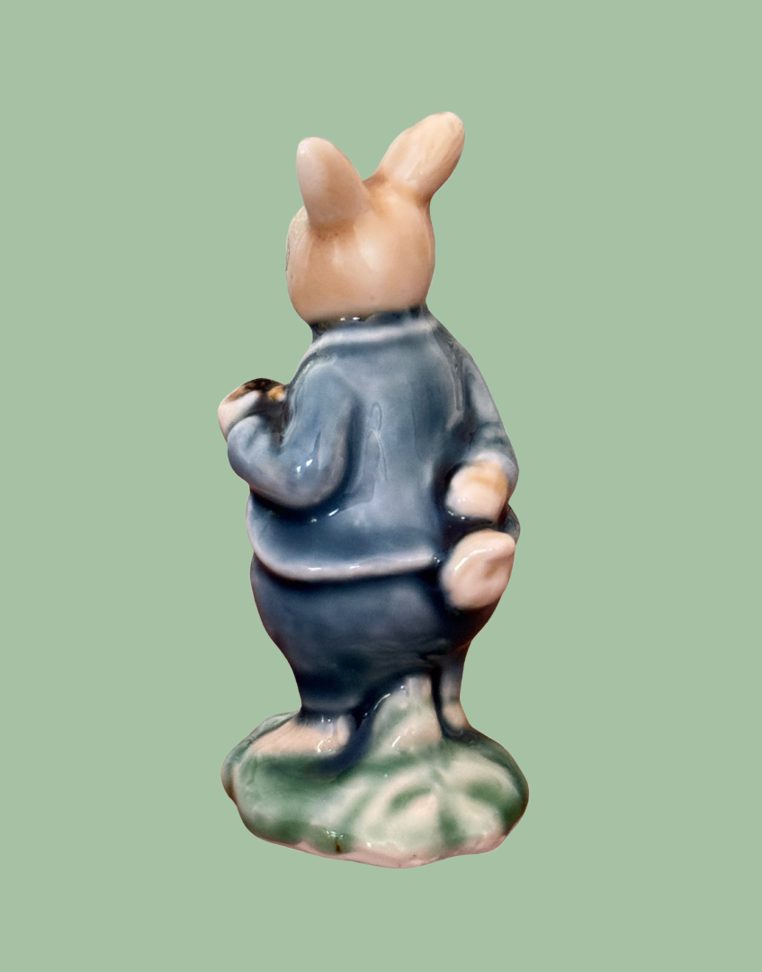 A ceramic figurine of a rabbit seen from the back, wearing a blue outfit and standing on a green base, set against a solid light green background.