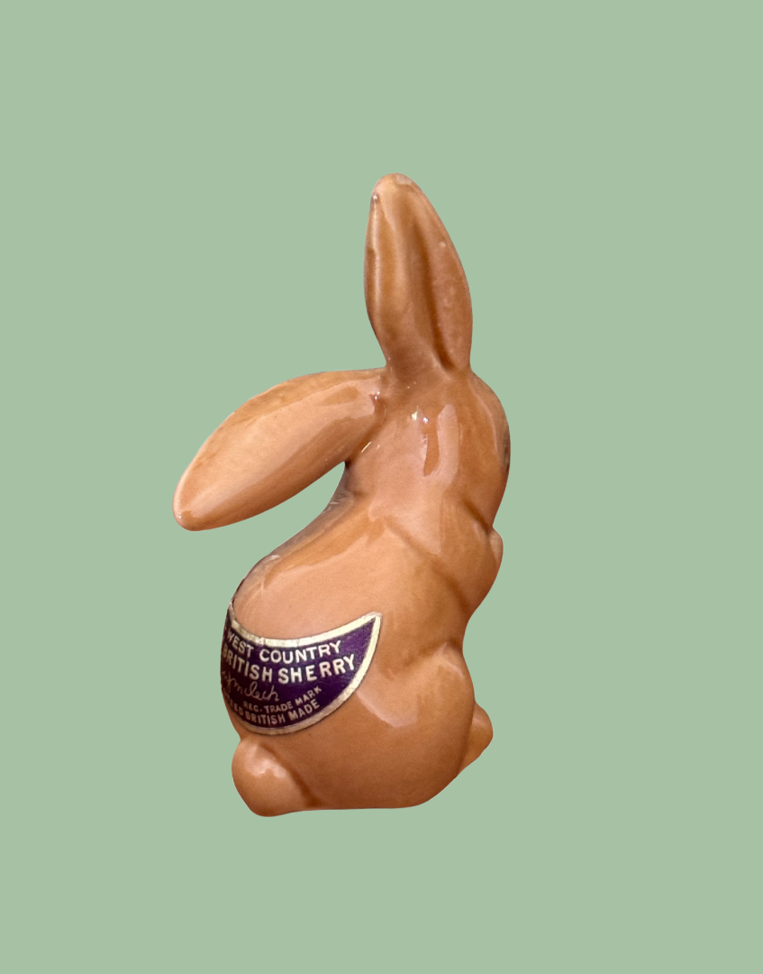 A glossy ceramic rabbit figurine in a warm tan-brown glaze, shown from the back against a solid sage green background. A purple and white label on its back reads 'COUNTRY BRITISH SHERRY'.