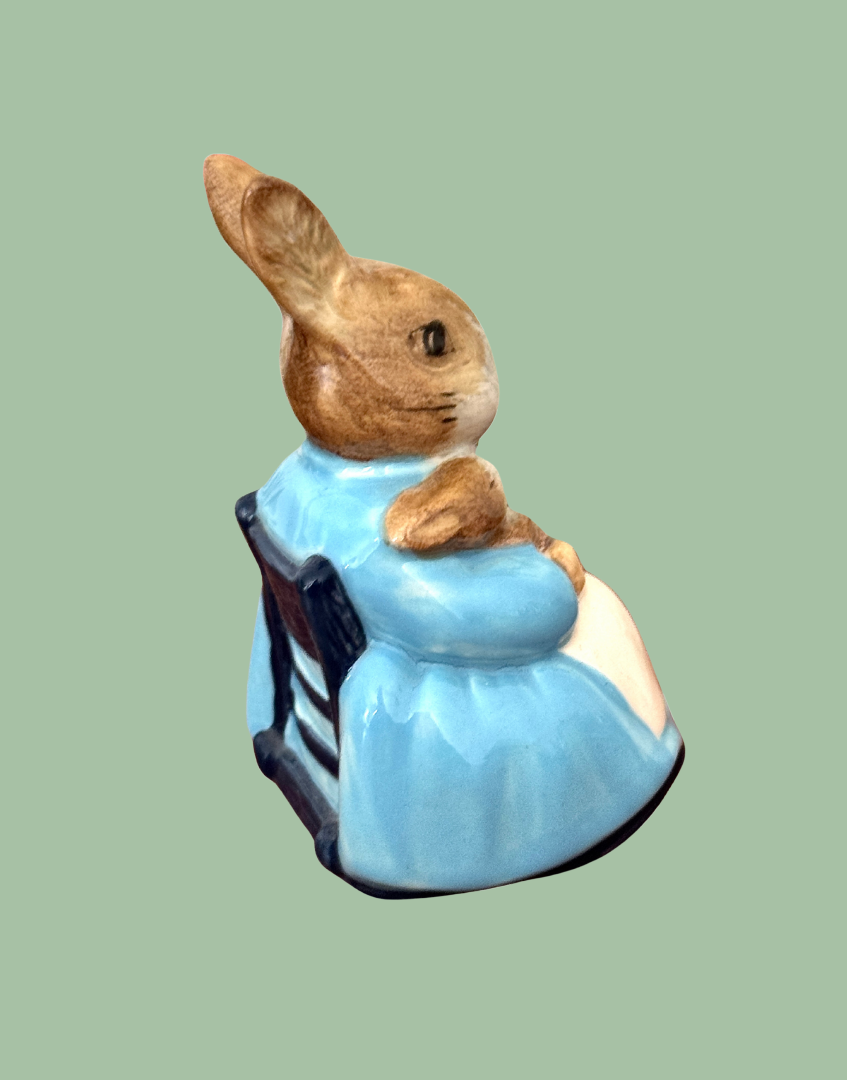 Back side view of Beatrix Potter seated rabbit figurine in blue dress and apron seated in a dark blue rocking chair
