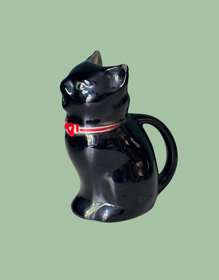 A glossy black cat-shaped ceramic pitcher with a red collar detail, set against a solid sage green background.