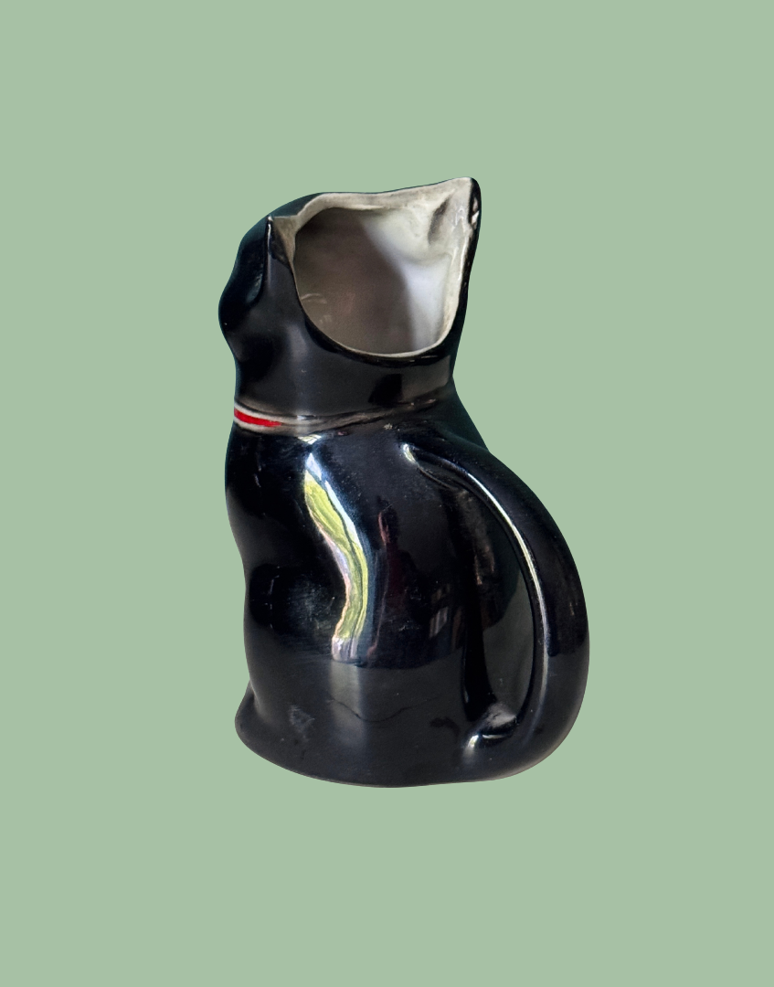A glossy black ceramic cat-shaped pitcher with a small red and white stripe detail on its neck, set against a solid sage green background.