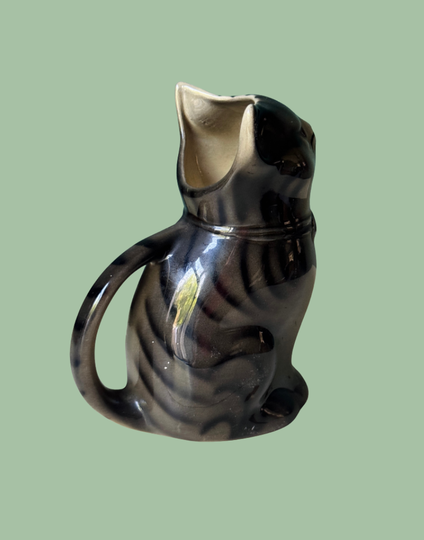 A glossy ceramic cat-shaped pitcher with a dark black and brown tabby pattern, set against a solid light green background.