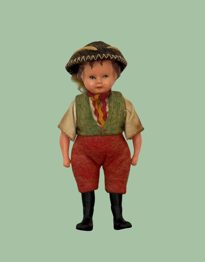 A vintage doll with a painted face and curly hair, wearing a brown patterned hat, a green vest, a red and yellow striped neckerchief, red pants, and black boots, set against a solid sage green background.