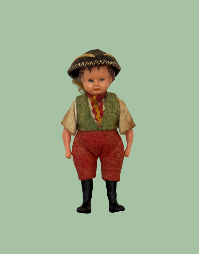 A vintage doll with a painted face and curly hair, wearing a brown patterned hat, a green vest, a red and yellow striped neckerchief, red pants, and black boots, set against a solid sage green background.