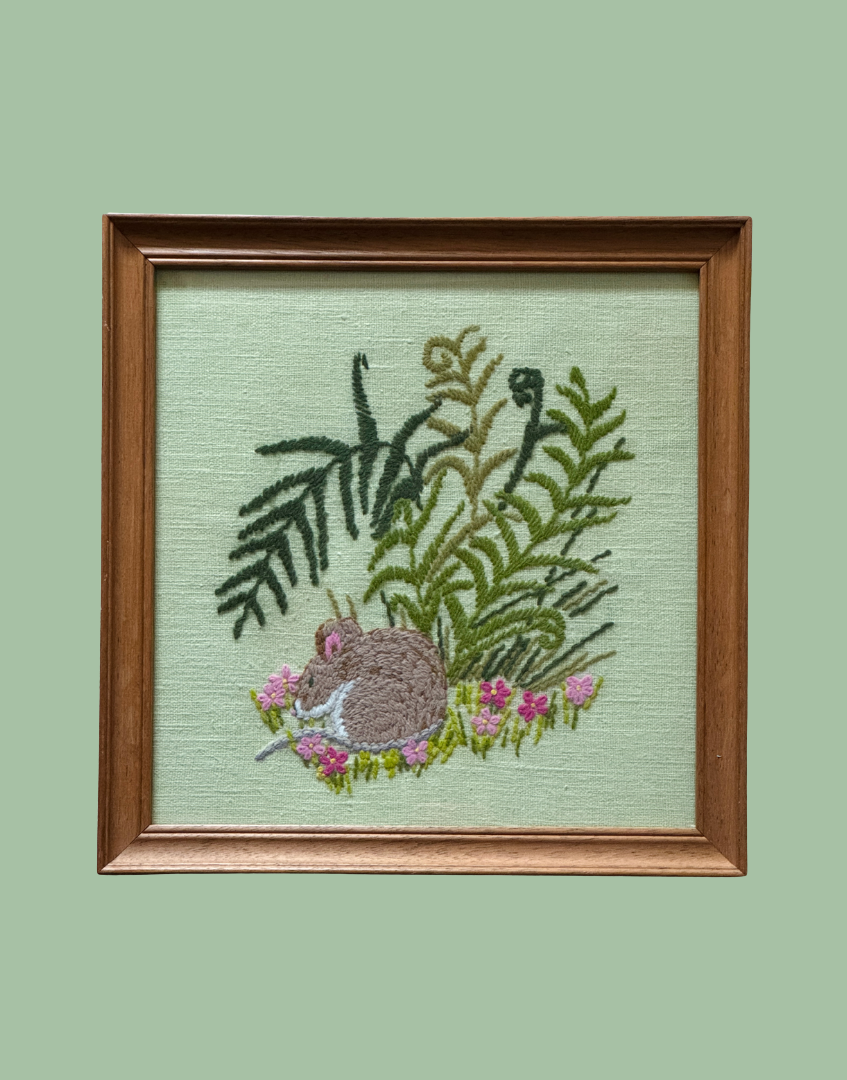 A framed embroidered artwork featuring a brown mouse among green ferns and pink flowers on a pale green fabric background, set in a medium brown wooden frame against a solid sage green backdrop.