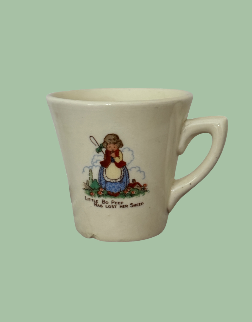 A vintage cream-colored ceramic coffee mug with a handle, featuring a printed illustration of a girl holding a pitchfork and the text 'Loving Bo Peep Has Lost Her Sheep' on a solid light green background.