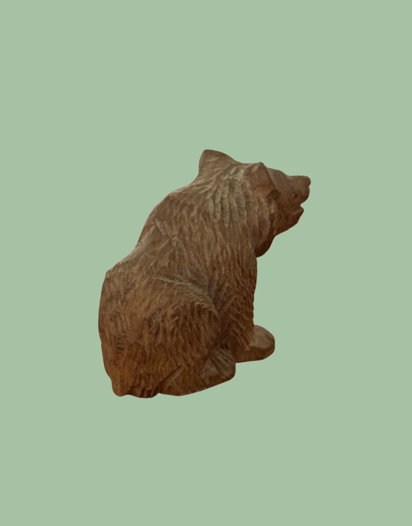 A carved wooden bear figurine shown from the back against a solid sage green background.