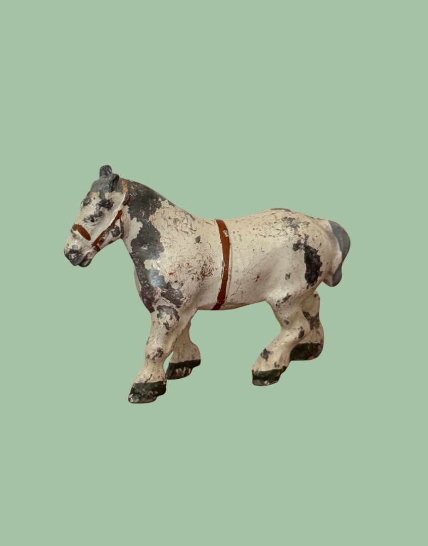 An antique cast iron toy horse figurine with a weathered white, black, and gray mottled finish and a brown harness, isolated on a solid sage green background.