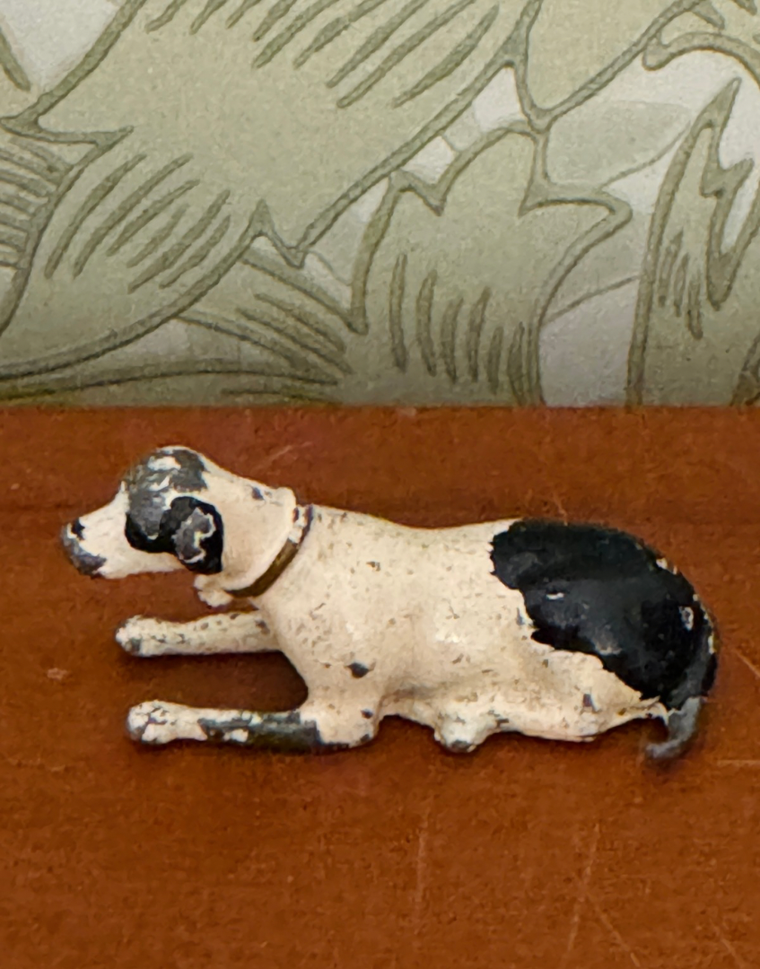 A small vintage-style painted dog figurine or toy lying down, with an off-white body and black patches, placed on a brown surface in front of a patterned wallpaper background.