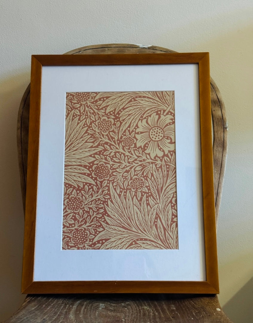 A framed botanical art print with a terracotta red pattern on a warm beige background, matted in white and enclosed in a medium brown wooden frame, leaning against a rustic wooden chair.