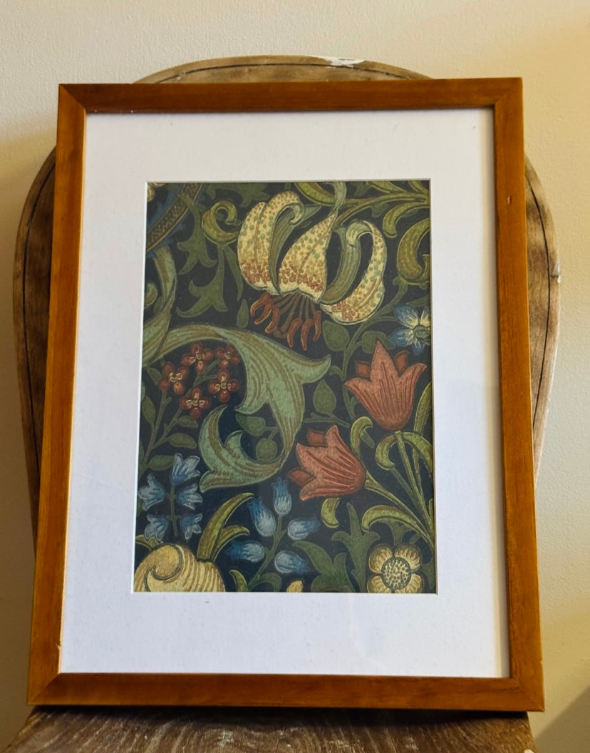 A framed botanical art print featuring a vintage-style floral pattern with green leaves, orange and blue flowers, and cream lilies on a dark background, housed in a warm brown wooden frame with a white mat.