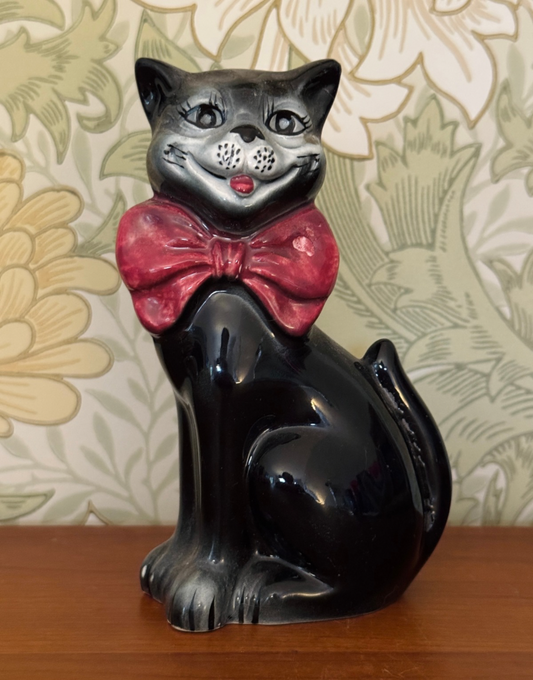 A glossy black cat figurine wearing a shiny red bow tie, set against a solid sage green background.