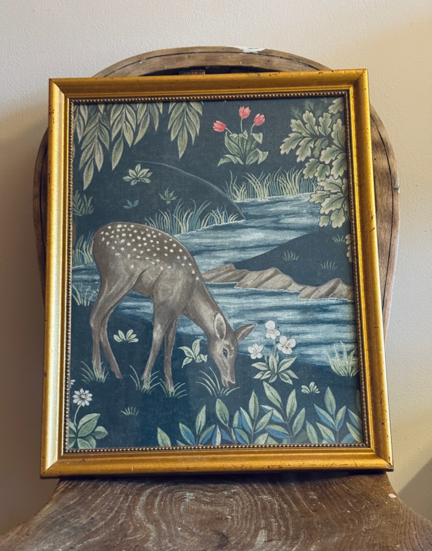 A framed William Morris art print depicting a spotted deer grazing by a river, with a dark blue background and green foliage, enclosed in a gold frame.