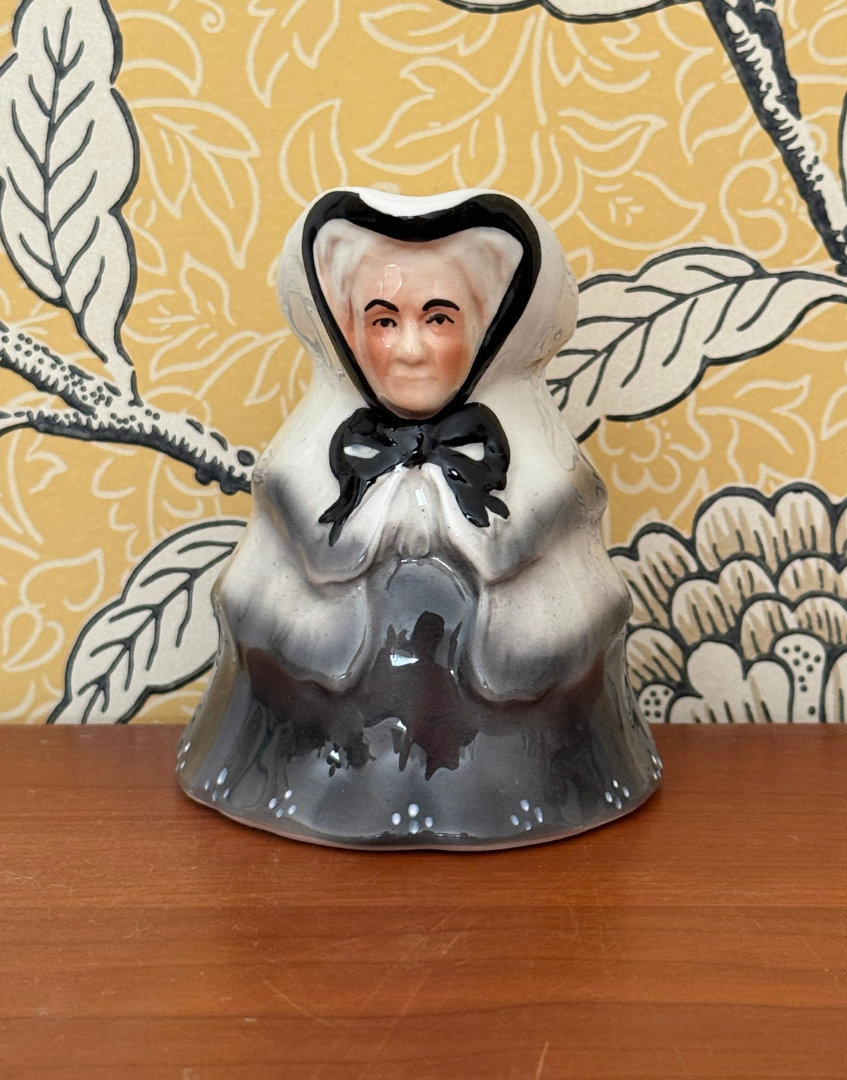 Tony Wood Little Old Lady creamer on a wooden surface against yellow botanical wallpaper, showing her black bow and grey skirt.